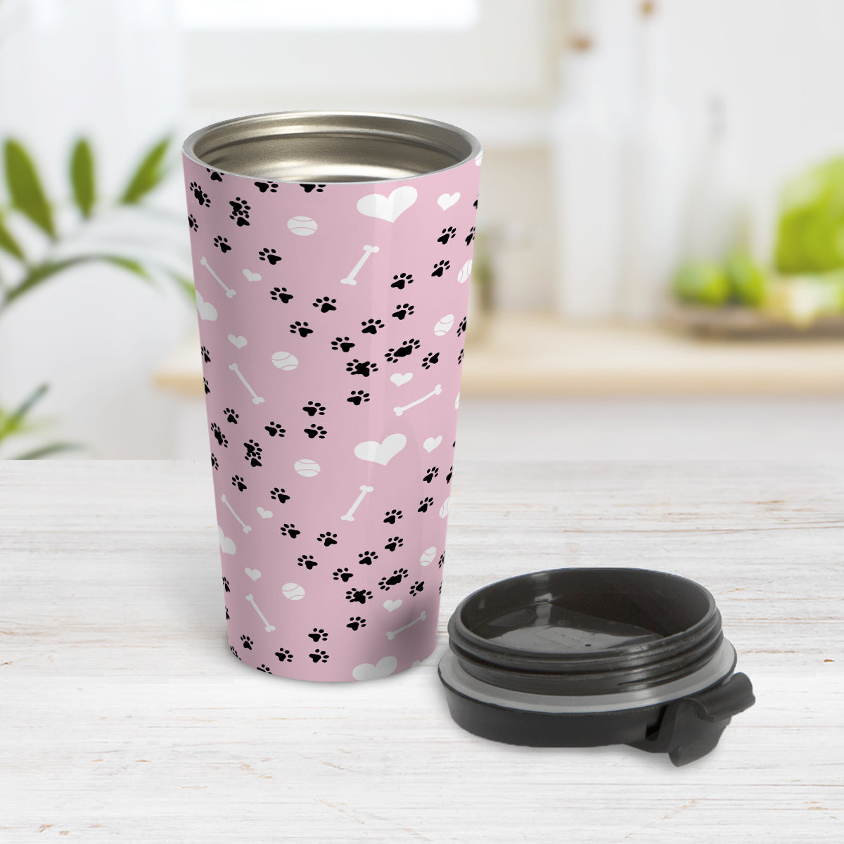 Puppy Run Pink Dog Travel Mug (15oz) at Amy's Coffee Mugs. A stainless steel travel mug designed with a pattern of chaotic tracks of puppy paw prints running around the mug with white dog bones, balls, and hearts over a light pink background. This mug is perfect for people who love the chaotic play of puppies, love dogs, and like pink travel mugs. Photo shows the open travel mug with the lid on the table beside it.