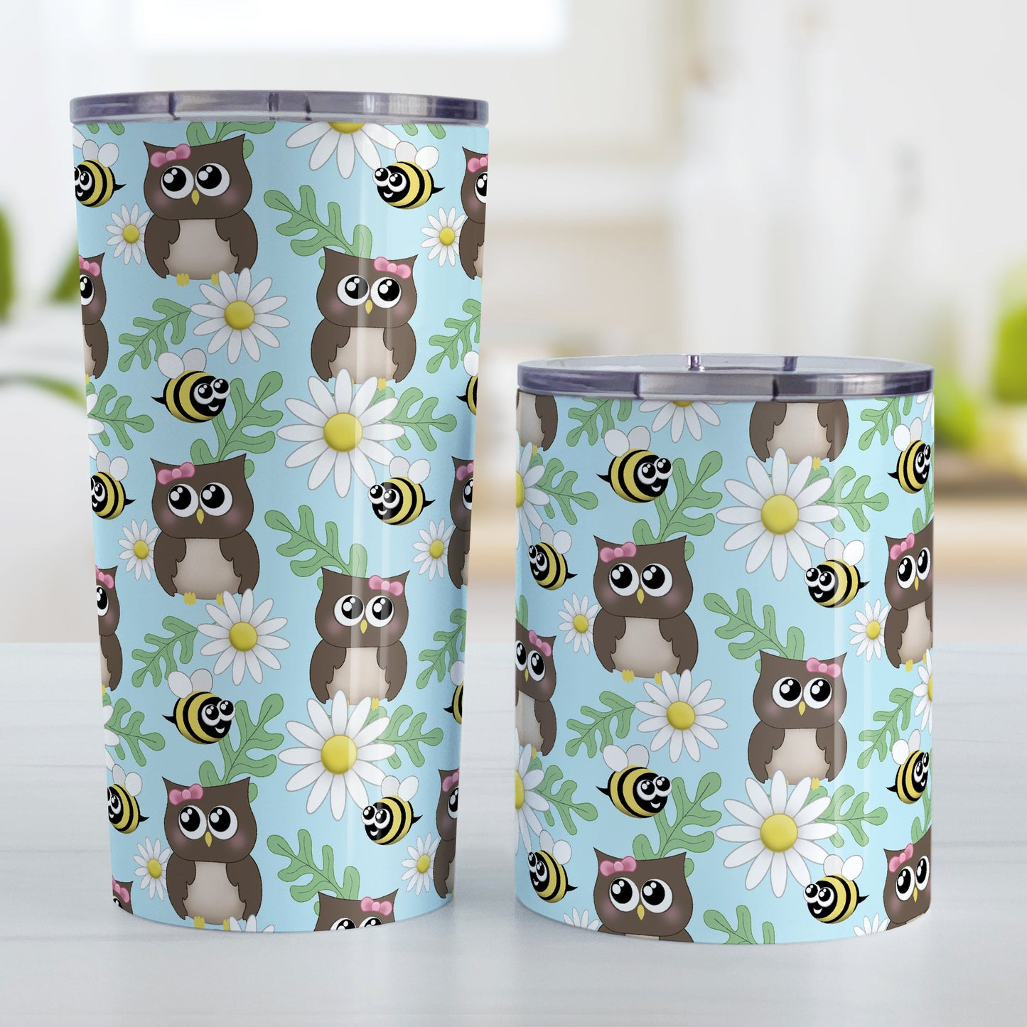 Spring Owl Bee Daisy Pattern Tumbler Cup (20oz and 10oz, stainless steel insulated) at Amy's Coffee Mugs. A tumbler cup designed with a pattern of brown owls with pink bows, happy bees, white daisy flowers, and green leaves over a light blue background color that wraps around the cup. Photo shows both sized cups on a table next to each other.