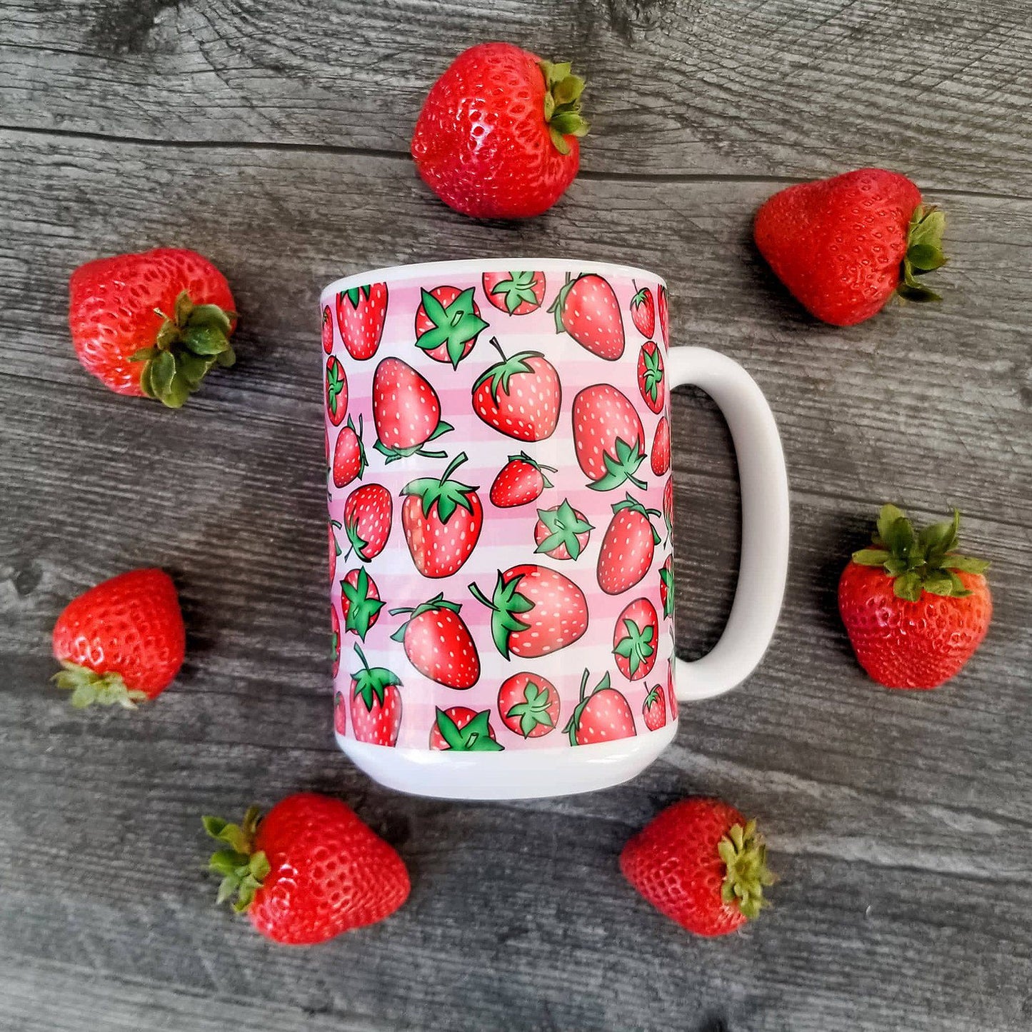 Strawberries on Pink Stripes Mug (15oz) in overhead view on weathered wood with strawberries around it. - Amy's Coffee Mugs. A ceramic coffee mug featuring a print of hand-drawn strawberries in red and green in an assorted pattern over a pink stripes background that wraps around the mug up to the handle.