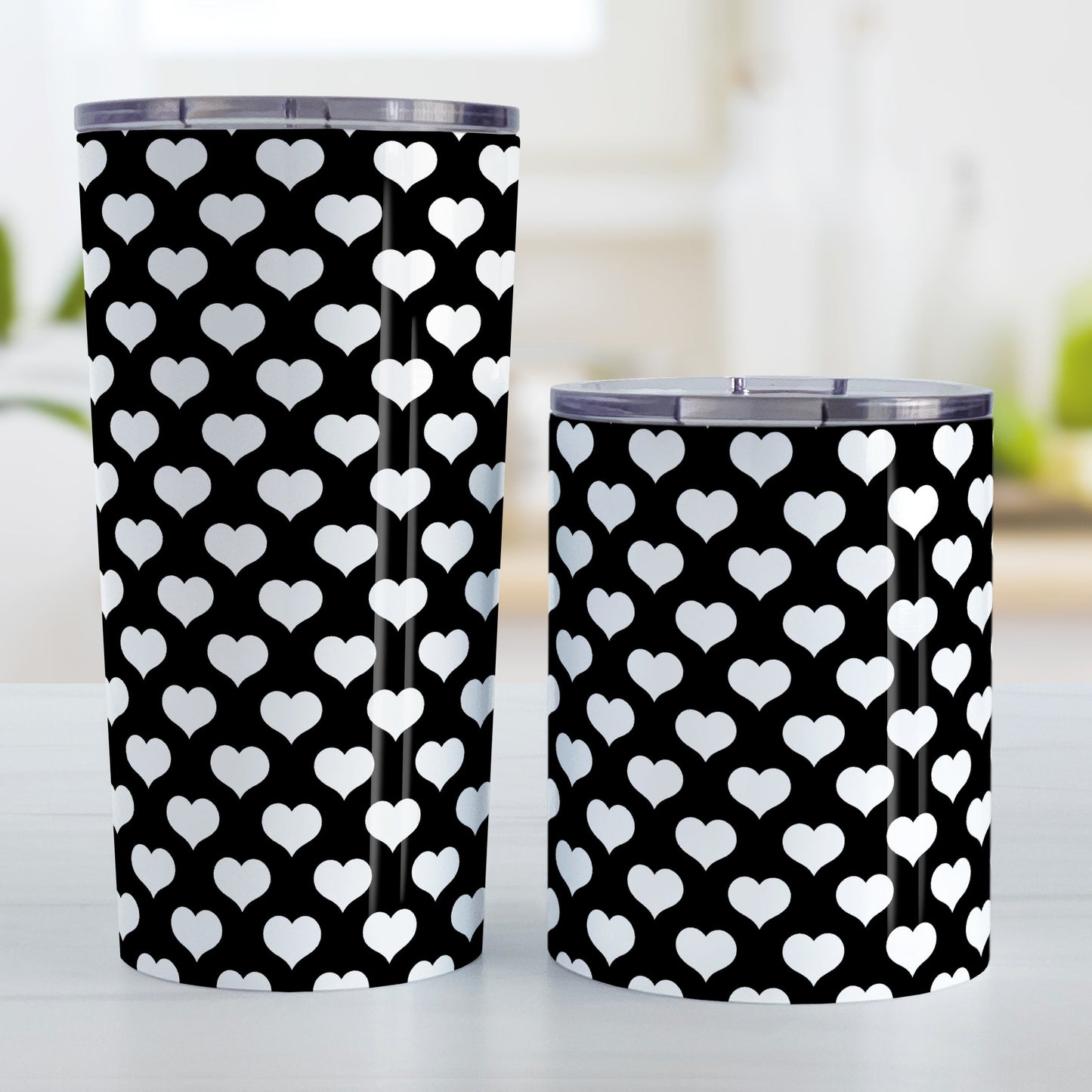 White Hearts Pattern Black Tumbler Cup (20oz and 10oz, stainless steel insulated) at Amy's Coffee Mugs. A tumbler cup designed with white hearts in a polka-dotted style pattern over a black background color that wraps around the cup. Photo shows both sized cups on a table next to each other.