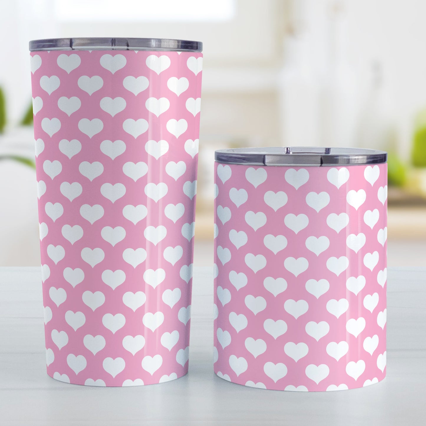 White Hearts Pattern Pink Tumbler Cup (20oz and 10oz, stainless steel insulated) at Amy's Coffee Mugs. A tumbler cup designed with white hearts in a polka-dotted style pattern over a pink background color that wraps around the cup. Photo shows both sized cups on a table next to each other.