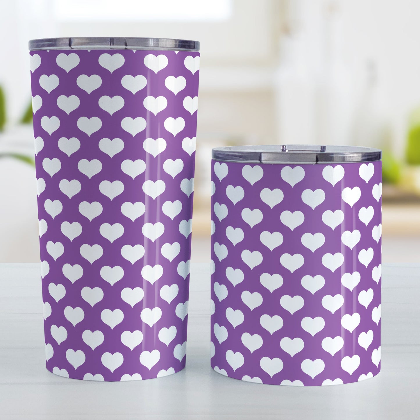 White Hearts Pattern Purple Tumbler Cup (20oz and 10oz, stainless steel insulated) at Amy's Coffee Mugs. A tumbler cup designed with white hearts in a polka-dotted style pattern over a purple background color that wraps around the cup. Photo shows both sized cups on a table next to each other.