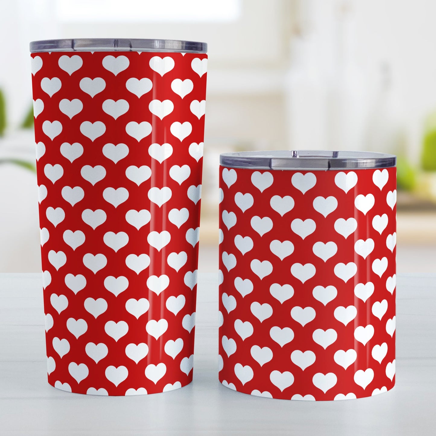 White Hearts Pattern Red Tumbler Cup (20oz and 10oz, stainless steel insulated) at Amy's Coffee Mugs. A tumbler cup designed with white hearts in a polka-dotted style pattern over a red background color that wraps around the cup. Photo shows both sized cups on a table next to each other.