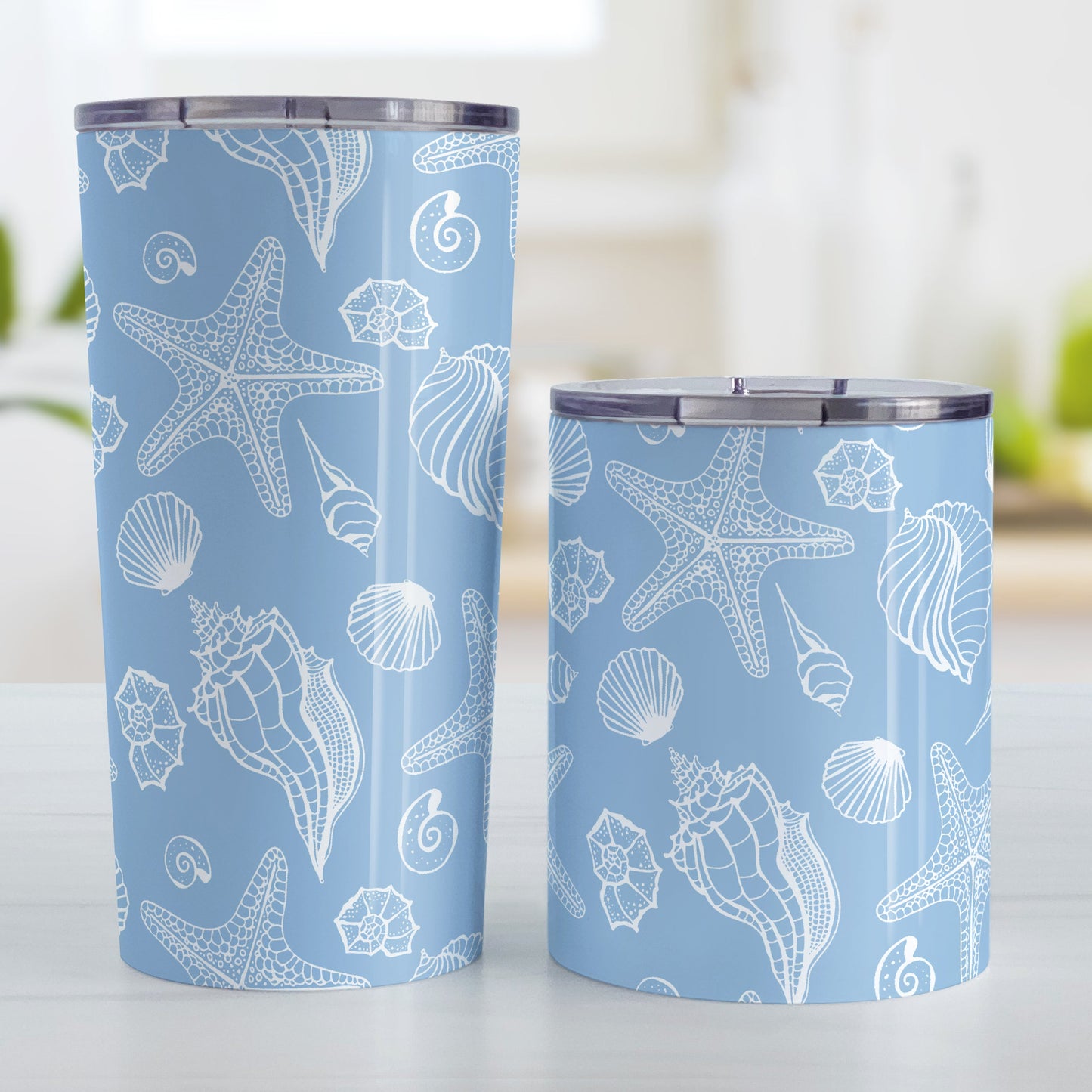 White Seashell Pattern Blue Beach Tumbler Cup (20oz and 10oz, stainless steel insulated) at Amy's Coffee Mugs. Tumbler Cups designed with a white seashell line drawing pattern over a blue background that wraps around the cups. Photo shows both sized cups on a table next to each other.