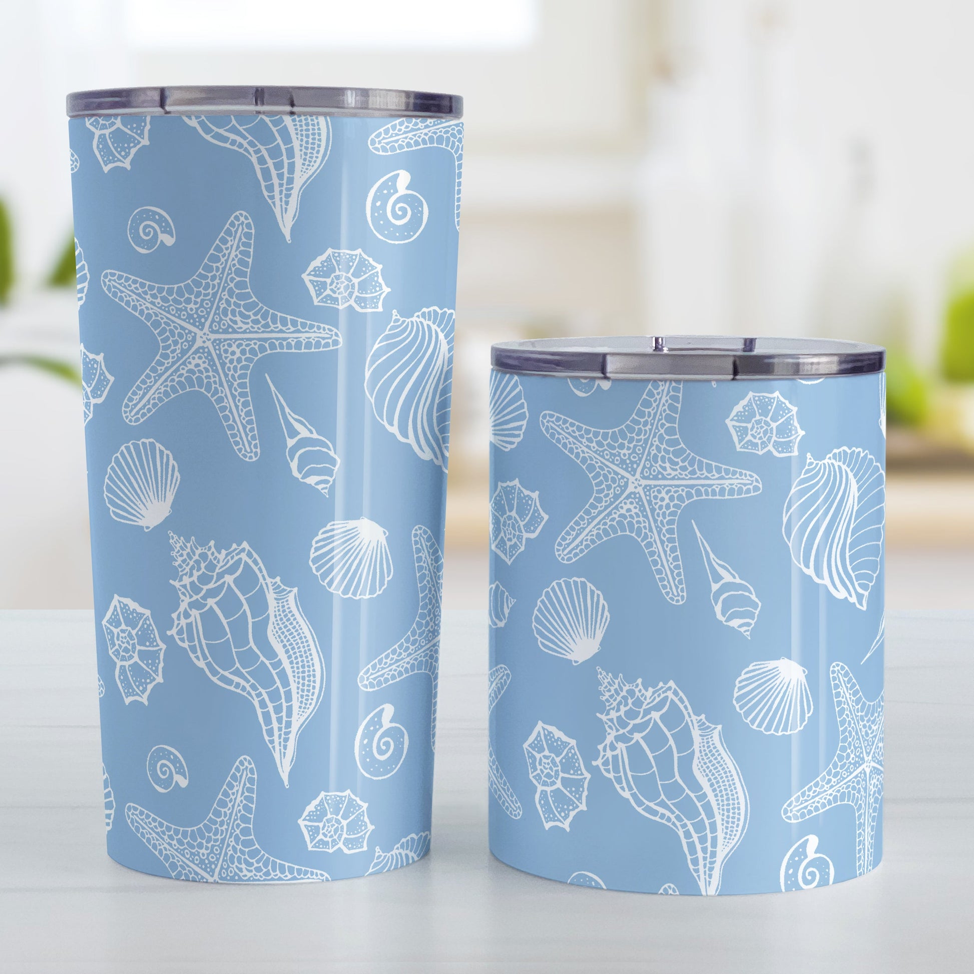 White Seashell Pattern Blue Beach Tumbler Cup (20oz and 10oz, stainless steel insulated) at Amy's Coffee Mugs. Tumbler Cups designed with a white seashell line drawing pattern over a blue background that wraps around the cups. Photo shows both sized cups on a table next to each other.