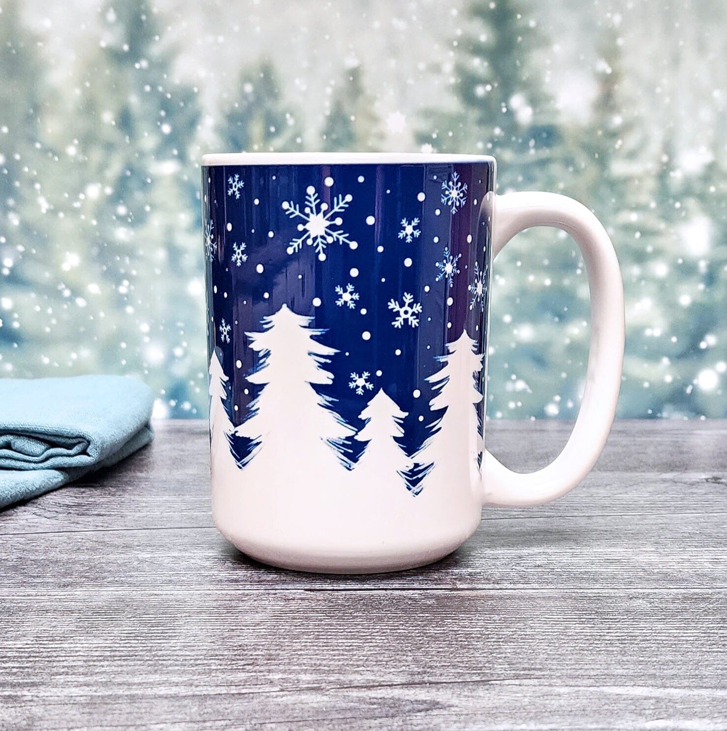 Winter Night Snow Mug (15oz) on a festive snowy tree background, from Amy's Coffee Mugs. A ceramic coffee mug designed with an illustrated winter night snow treeline. The design features a navy blue sky filled with white snowflakes over a row of white pine trees along the bottom.