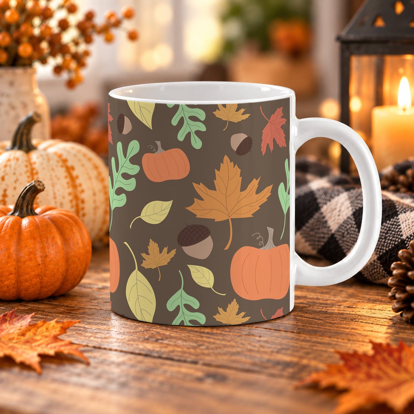 An 11 oz white ceramic mug with a dark brown autumn-themed pattern featuring pumpkins, maple leaves, acorns, and fall leaves, styled on a wooden table with pumpkins, a candle, and a plaid cloth.