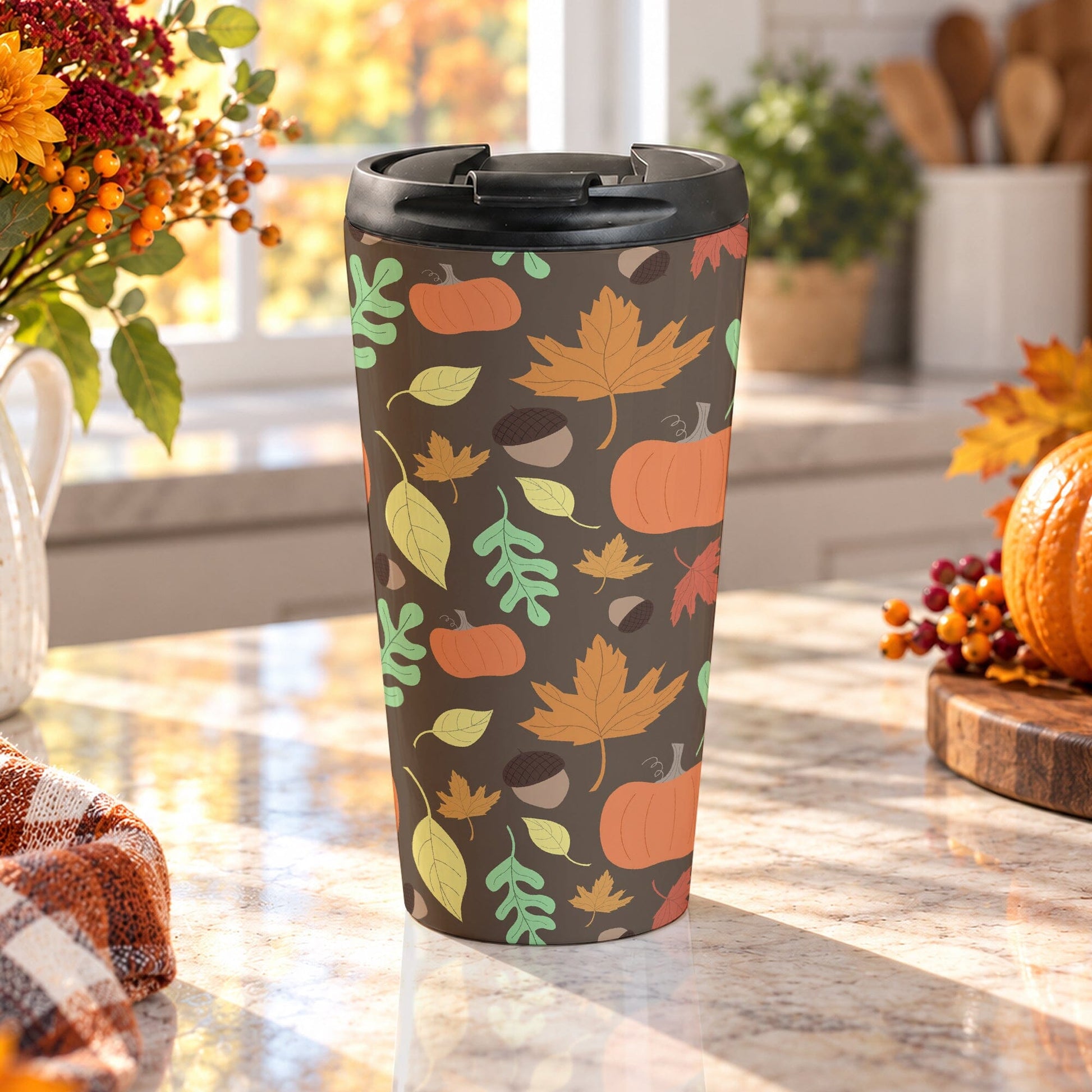 15 oz fall travel mug with orange pumpkins, acorns, and autumn leaves in orange, yellow, green, and red over a brown background, on a granite countertop in a kitchen filled with fall decor, including autumn plants, a pumpkin, and an orange plaid tea towel.