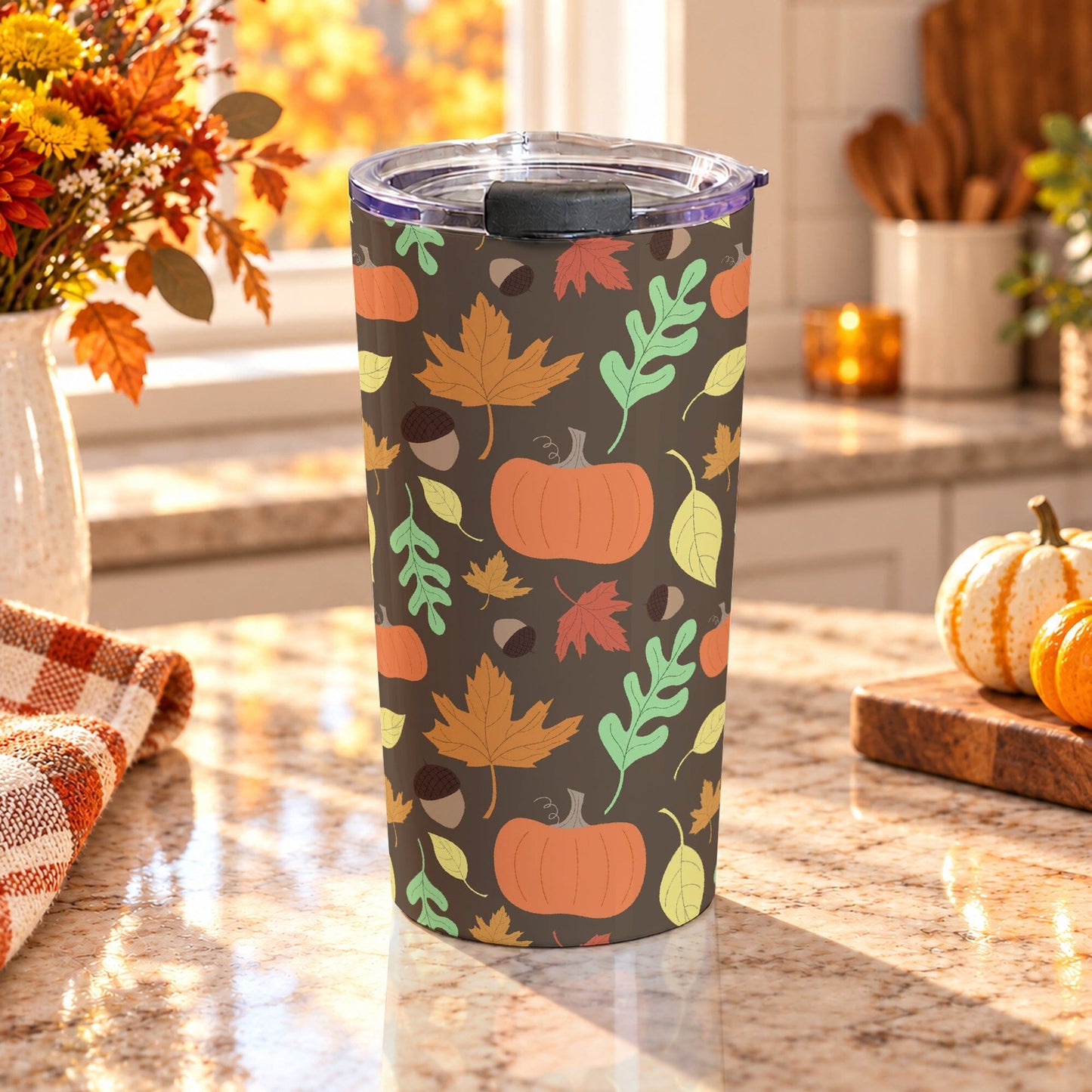 20 oz fall tumbler cup with orange pumpkins, acorns, and orange, yellow, green, and red autumn leaves over a brown background, on a granite countertop in a kitchen with fall decor including autumn plants, pumpkins, and an orange plaid tea towel.