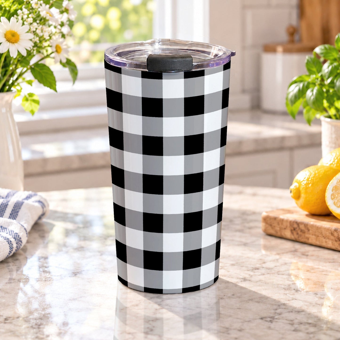 Black and white buffalo plaid tumbler cup with gray accents, large checked pattern, and a 20 oz insulated stainless steel body, on a granite countertop in a bright kitchen with spring/summer flowers, green plants, a tea towel, and lemons on a wood cutting board.