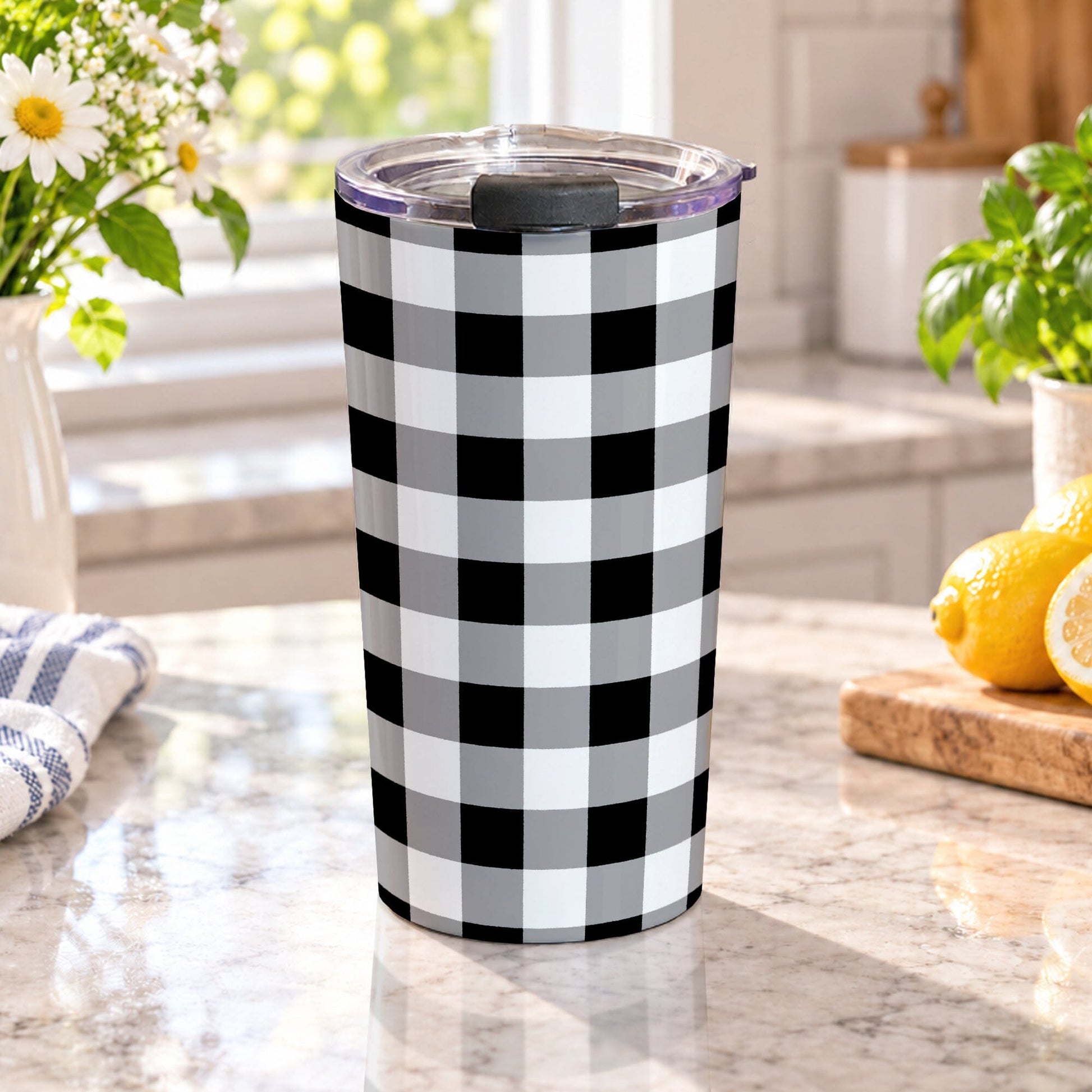 Black and white buffalo plaid tumbler cup with gray accents, large checked pattern, and a 20 oz insulated stainless steel body, on a granite countertop in a bright kitchen with spring/summer flowers, green plants, a tea towel, and lemons on a wood cutting board.