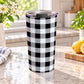 Black and white buffalo plaid tumbler cup with gray accents, large checked pattern, and a 20 oz insulated stainless steel body, on a granite countertop in a bright kitchen with spring/summer flowers, green plants, a tea towel, and lemons on a wood cutting board.