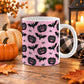 White 11 oz ceramic Halloween mug with black jack-o'-lanterns, bats, spiders, and white spider webs on a pink background, sitting upon a wooden table with fall decor around it.