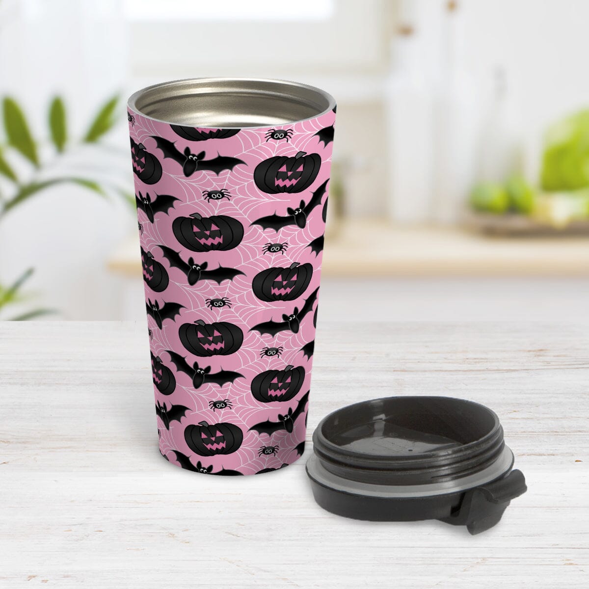 Black Pumpkins Bats and Spiders Pink Halloween Travel Mug (15oz) at Amy's Coffee Mugs. A travel mug designed with a pattern of black carved pumpkins, bats, and spiders in their webs in a pattern over a pink background color that wraps around the mug. Photo shows the mug on a table with its lid beside it.