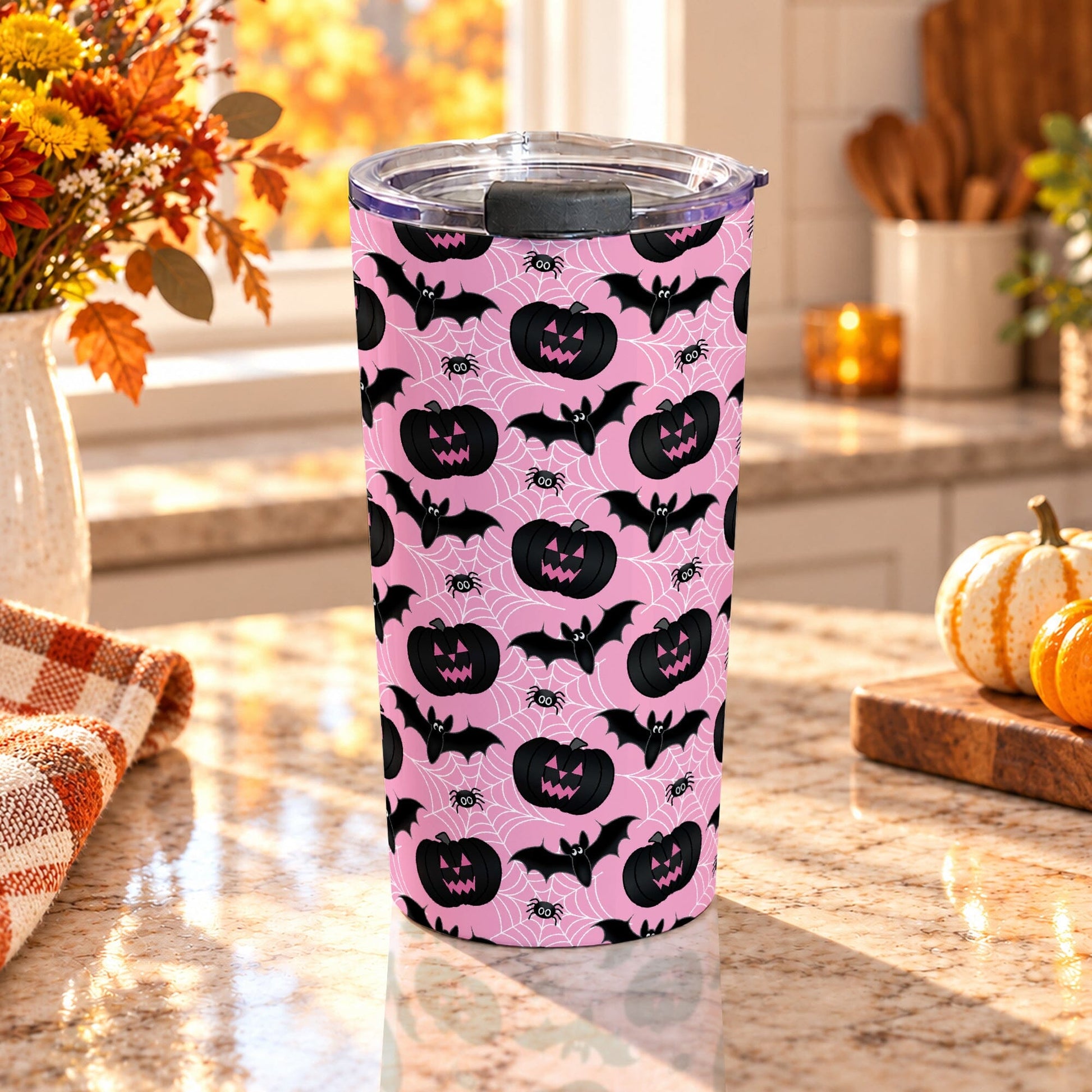 20 oz insulated stainless steel Halloween tumbler cup with black jack-o'-lanterns, bats, spiders, and white spider webs on a pink background, on a granite countertop in a kitchen with fall decor including autumn plants, pumpkins, and an orange plaid tea towel.