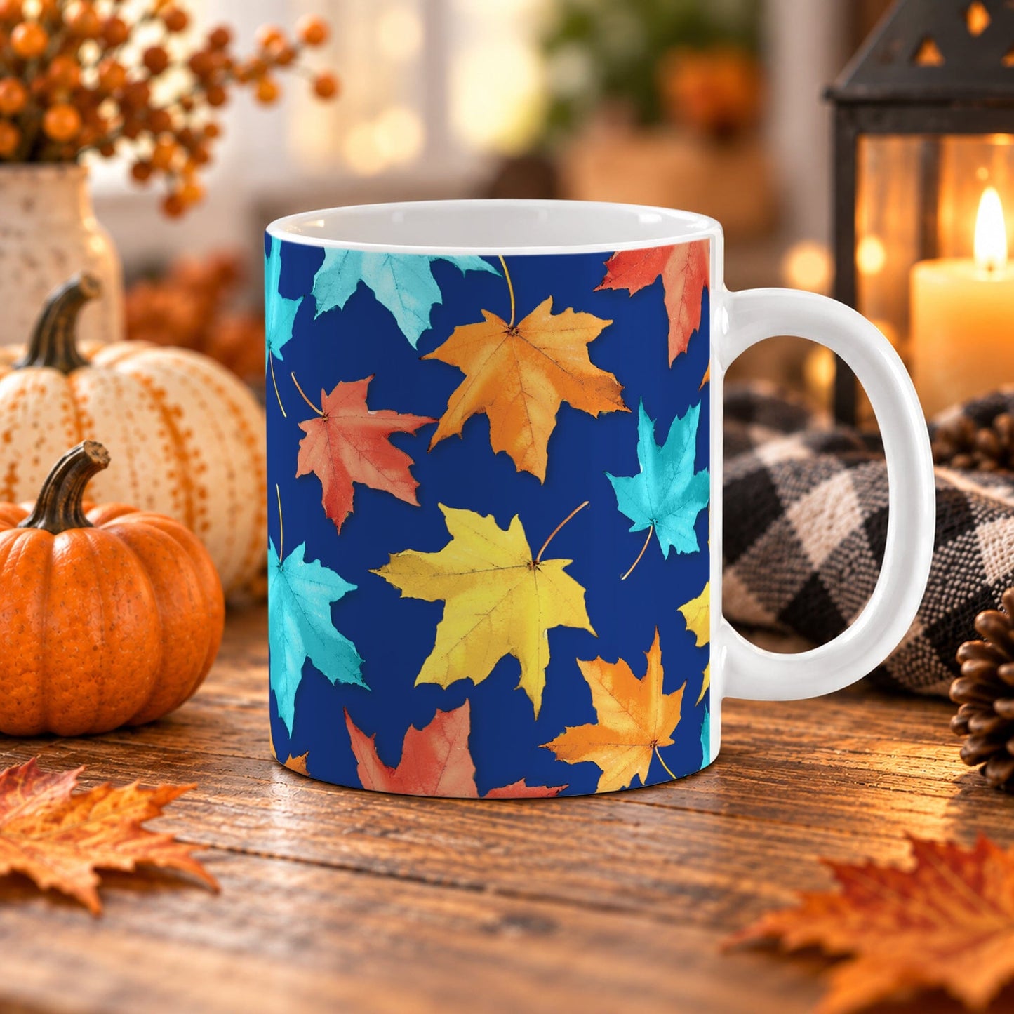 11 oz Blue Fall Leaves Mug with colorful turquoise, orange, and yellow autumn leaves over a deep navy blue background, on a wooden table with fall decor around it including pumpkins, autumn leaves, a candle lantern, and a cozy checkered blanket.