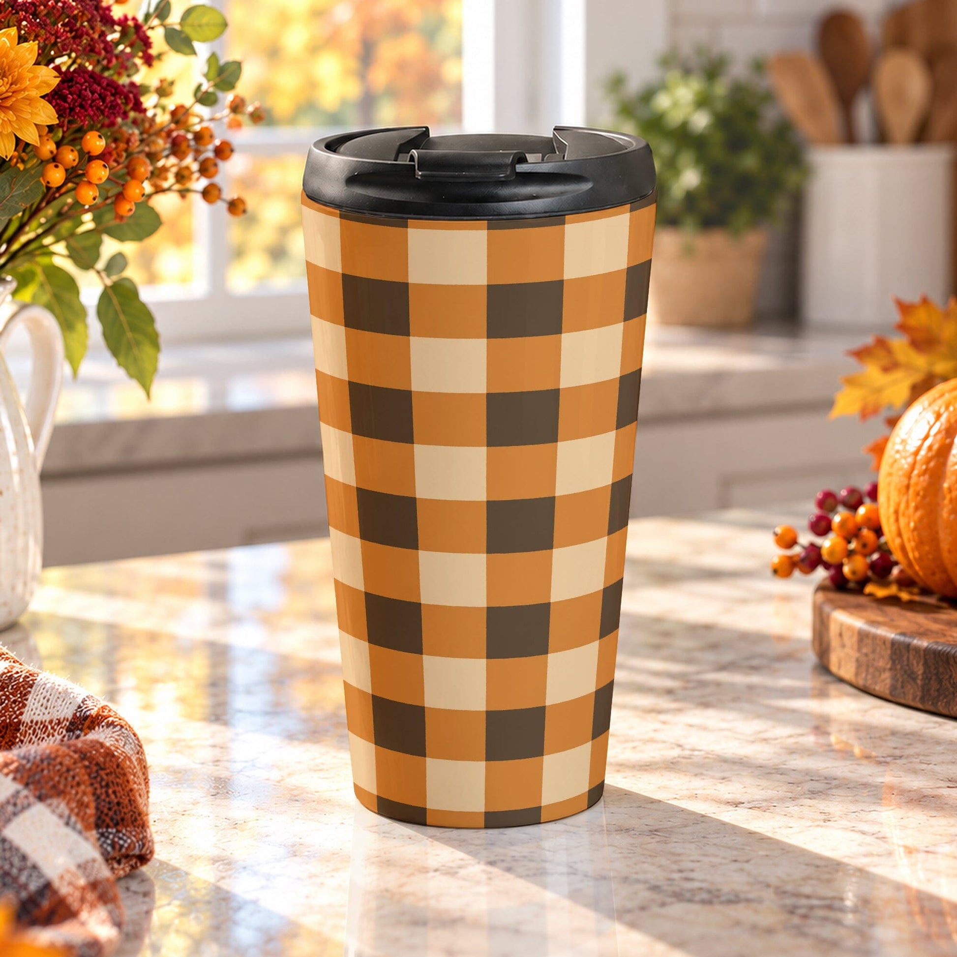 15 oz brown and orange buffalo plaid travel mug with brown, orange, and beige buffalo check pattern, on a granite countertop in a kitchen with fall decor including autumn plants, a pumpkin, and an orange plaid tea towel.