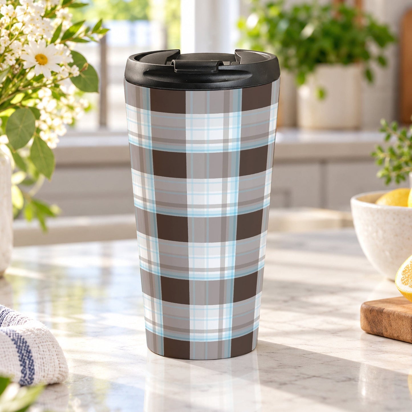 15 oz brown and blue plaid travel mug with dark brown, light brown, white, and soft blue layered plaid pattern, on a granite countertop in a bright kitchen with spring/summer daisies, green plants, tea towel, and lemons on a cutting board.