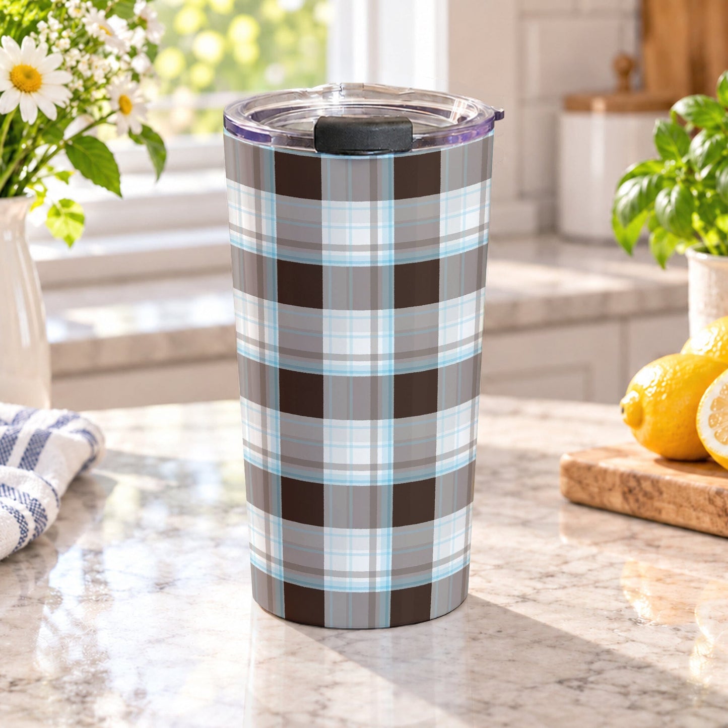 20 oz brown and blue plaid tumbler cup with brown, taupe, white, and light blue plaid pattern, on a granite countertop in a bright kitchen with spring/summer daisies, green plants, tea towel, and lemons on a wood cutting board.