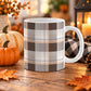 11 oz brown orange plaid mug with dark brown, light brown, white, and warm orange layered plaid pattern, on a wooden table with fall decor around it, including pumpkins, autumn leaves, a candle lantern, and a cozy checkered blanket.