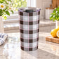 20 oz brown and pink plaid tumbler cup with a brown, taupe, white, and soft pink pattern, on a granite countertop in a bright kitchen with spring/summer daisies, green plants, tea towel, and lemons on a wood cutting board.