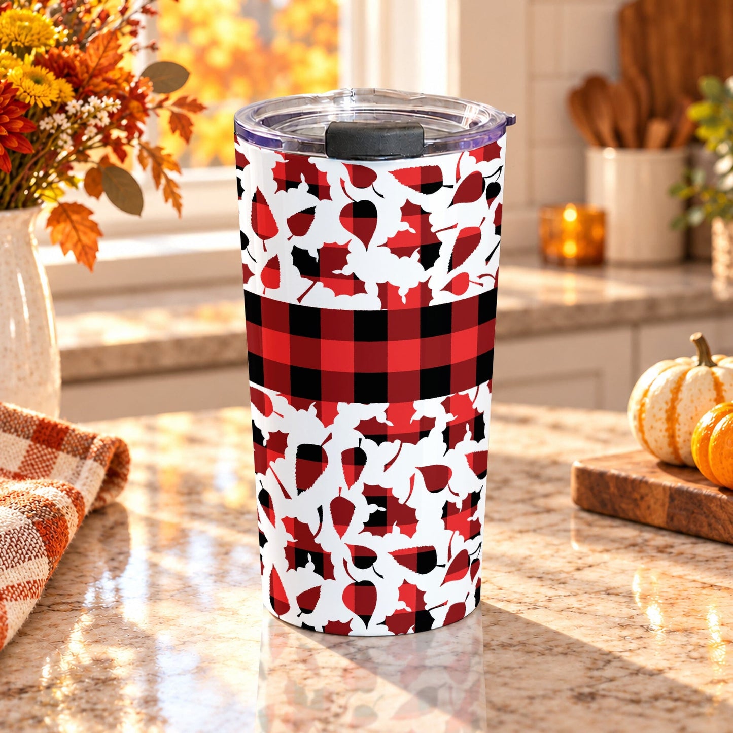 20 oz fall tumbler cup with red and black buffalo plaid, assorted autumn leaf shapes, and a wide checkered band around the center, on a granite countertop in a kitchen with fall decor including autumn plants, pumpkins, and an orange plaid tea towel.