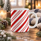 An 11 oz white ceramic mug with a red and white diagonal candy cane stripe pattern, set on a rustic wooden table surrounded by holiday decorations like pinecones, evergreen branches, and a knitted blanket.