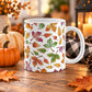 Fall 11 oz ceramic coffee mug with colorful red, orange, yellow, green, purple, and brown autumn leaves on a white background, shown on a wooden table with fall decor.