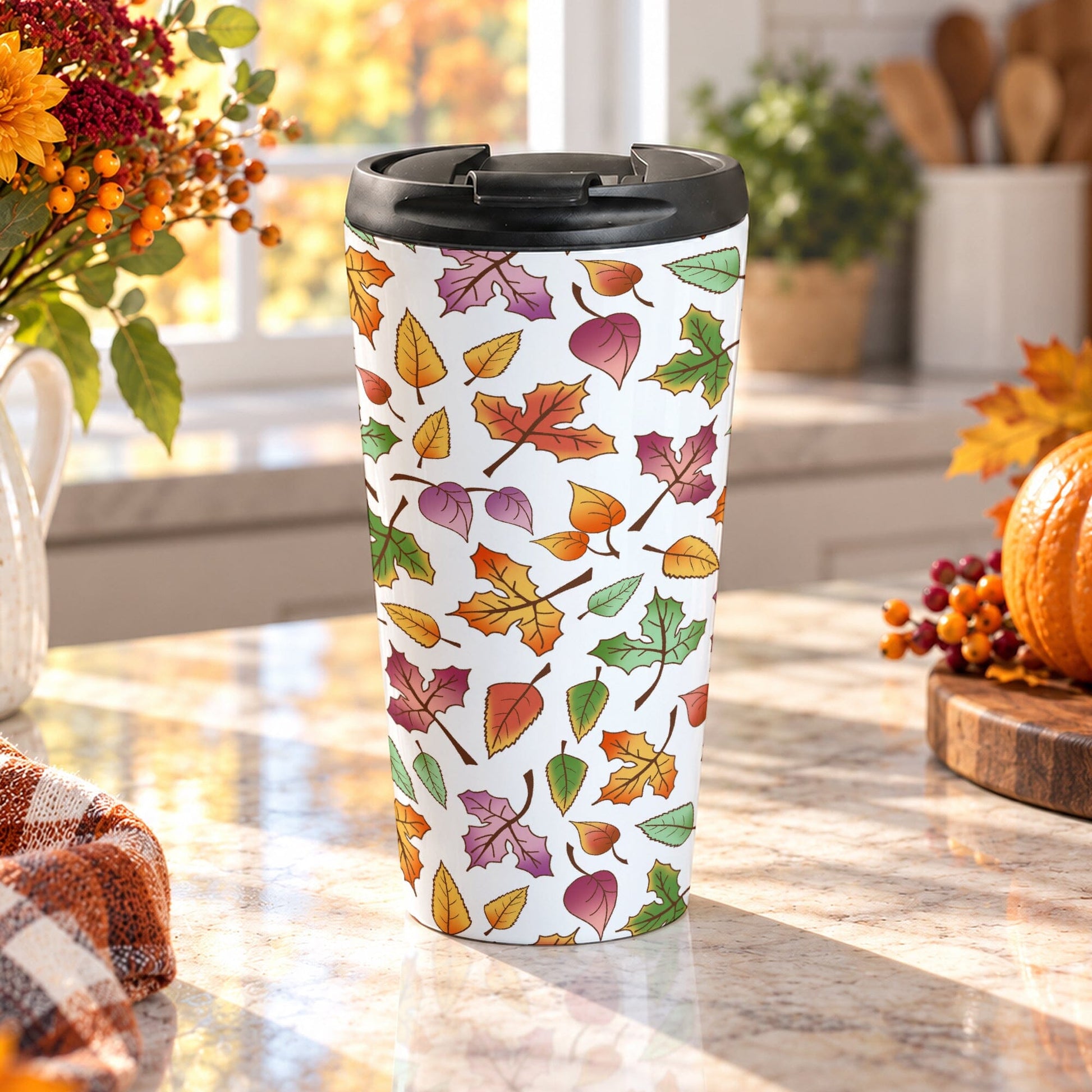 15 oz Changing Leaves Fall Travel Mug with colorful orange, yellow, red, green, and purple autumn leaves, sitting on a granite countertop in a fall-themed kitchen with autumn plants, pumpkin, and an orange plaid tea towel.