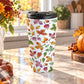 15 oz Changing Leaves Fall Travel Mug with colorful orange, yellow, red, green, and purple autumn leaves, sitting on a granite countertop in a fall-themed kitchen with autumn plants, pumpkin, and an orange plaid tea towel.