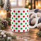 11 oz Christmas mug with red and green polka dot pattern on white ceramic, on a wooden table with winter decor around it including pine cones, pine boughs, candles, and a cozy plaid blanket.