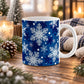 11 oz blue snowflake winter mug with large white and light blue snowflakes, dark blue accents, and smaller snowflakes on a classic blue background, shown on a wooden table with winter decor around it.