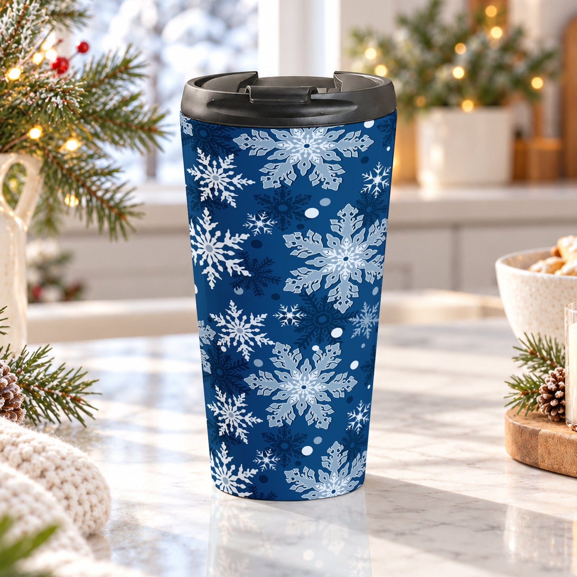 15 oz classic blue snowflake winter travel mug with white, light blue, and dark blue snowflakes, on a granite countertop in a kitchen with cozy winter decor and small warm white Christmas lights.