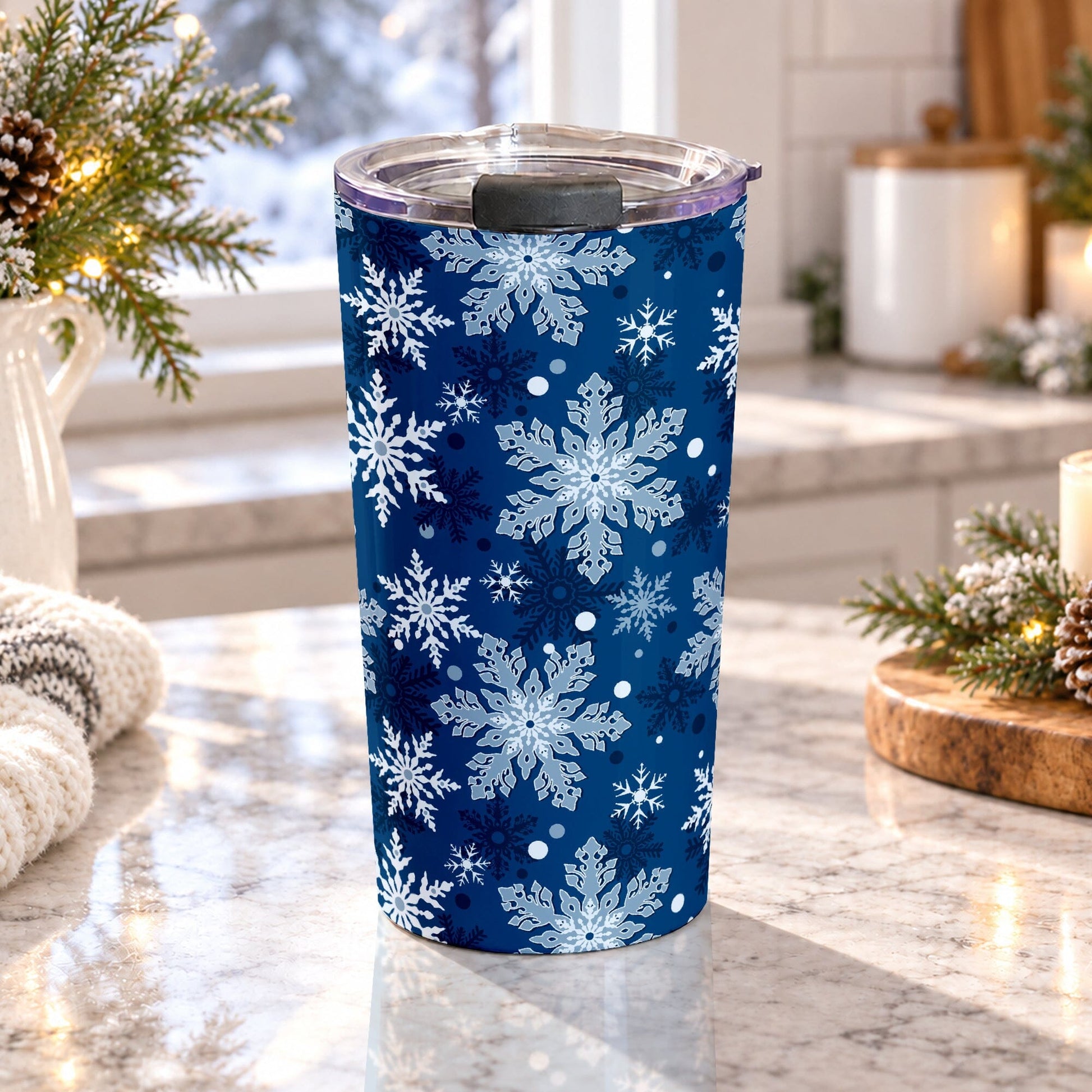 A 20 oz tumbler cup designed with a pattern of different shades of blue snowflakes over a classic blue background color that wraps around the cup, on a granite countertop in a kitchen with light rustic winter decor including pine cones, pine boughs, and a cozy blanket.