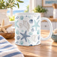 11 oz coastal coffee mug with navy, aqua, and beige seashells, scallop shells, spiral shells, and starfish on white ceramic, set on a wooden table in a bright, coastal-themed kitchen.