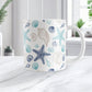 White mug with blue and beige seashell pattern on a white marble surface.