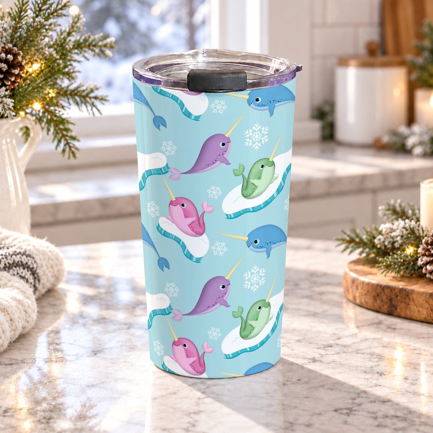 Colorful narwhal tumbler cup with pink, purple, blue and green narwhals, snowflakes, icy shapes, and a 20 oz insulated stainless steel body, on a granite countertop in a bright kitchen with light rustic decor including pine cones, pine boughs, lights, and a cozy blanket.