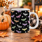 White 11 oz ceramic Halloween mug with colorful orange, purple, pink, green and silver bats, white stars and dots on a black background. The mug sits on a wooden table with fall decor around it.