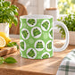 A white ceramic mug with a green and white frog pattern, set on a wooden table in a bright spring kitchen environment with a lemon, basil, and plants in the background.