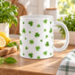 11 oz St. Patrick’s Day mug with small green shamrocks and four-leaf clovers scattered across a white ceramic background, shown on a wooden tabletop in a kitchen with spring plants and decor.