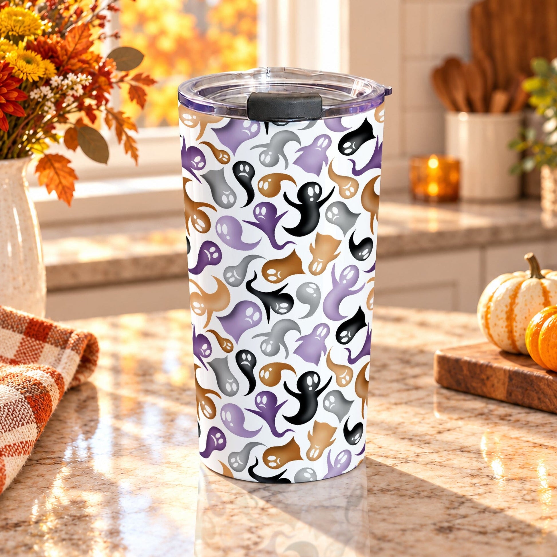 20 oz insulated stainless steel Halloween tumbler cup with purple, orange, black, and gray ghosts and spirits on a white background, on a granite countertop in a kitchen with fall decor including autumn plants, pumpkins, and an orange plaid tea towel.