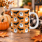 White 11 oz ceramic Halloween mug with white ghosts, black bats, spiders, stars and spider webs on an orange background, on a wooden table with autumn decor including pumpkins, autumn leaves, a candle lantern, and a cozy checkered blanket.