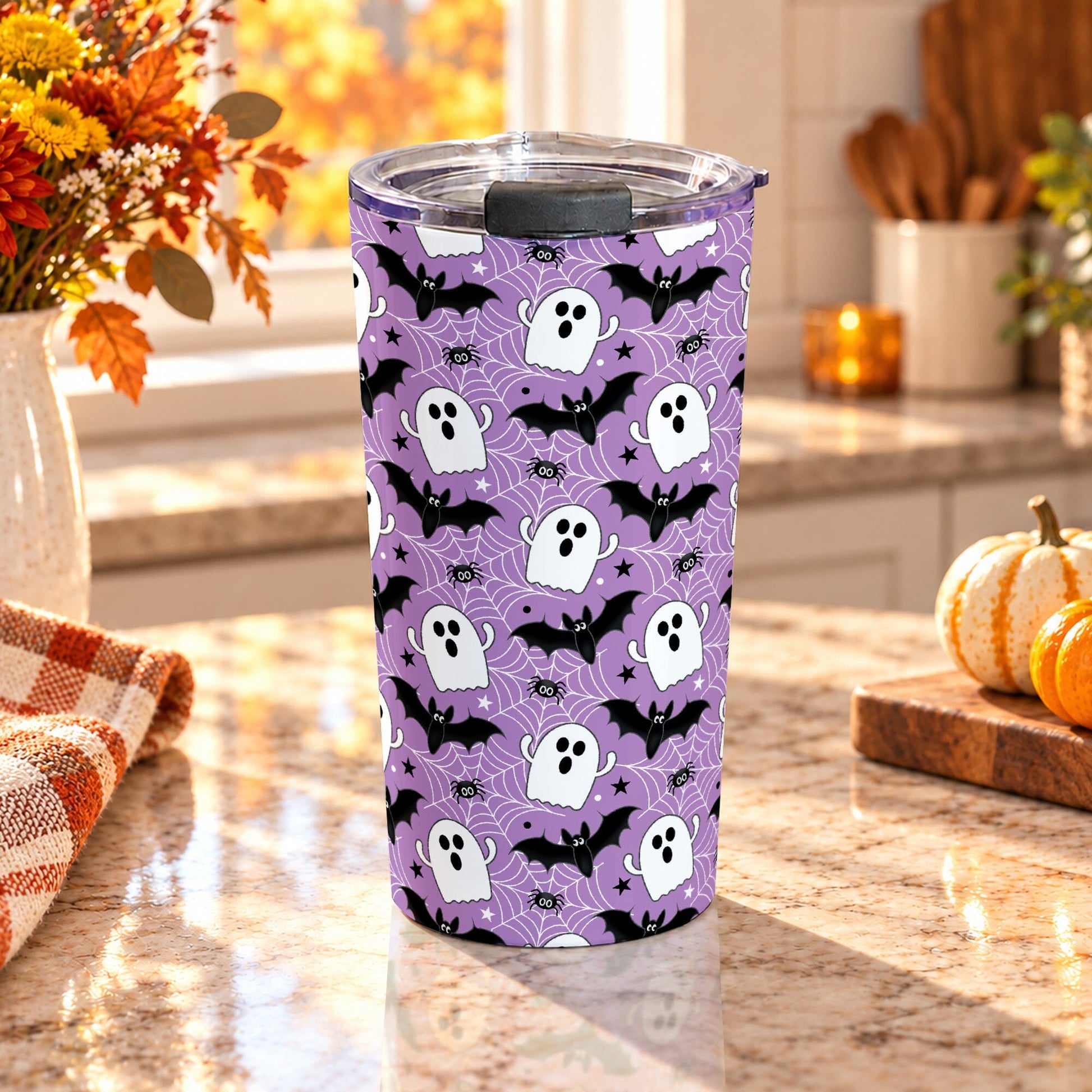 20 oz insulated stainless steel Halloween tumbler cup with white ghosts, black bats, spiders, stars, and white spider webs on a purple background, on a granite countertop in a kitchen with fall decor including autumn plants, pumpkins, and an orange plaid tea towel.