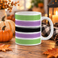 White 11 oz ceramic Halloween coffee mug with a print of horizontal green, purple and black painted brush strokes wrapping around the mug up to the handle, displayed on a wooden table with cozy fall decor around it.