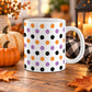 White 11 oz ceramic Halloween mug with repeating orange, purple and black polka dots, displayed on a wooden table with fall decor around it.