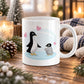 11 oz penguin mug with an adult black-and-white penguin facing a fluffy gray baby penguin, surrounded by pink hearts on a pale blue background design, displayed on a wooden table with winter decor around it.