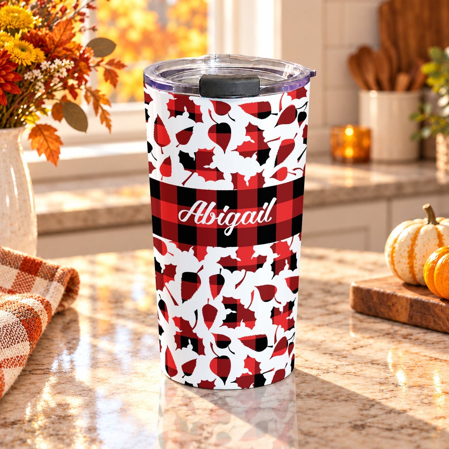 20 oz personalized fall tumbler cup with red and black buffalo plaid, assorted autumn leaf shapes, custom white script name, and wide checkered center band, on a granite countertop in a kitchen with fall decor including autumn plants, pumpkins, and an orange plaid tea towel.