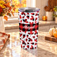 20 oz personalized fall tumbler cup with red and black buffalo plaid, assorted autumn leaf shapes, custom white script name, and wide checkered center band, on a granite countertop in a kitchen with fall decor including autumn plants, pumpkins, and an orange plaid tea towel.