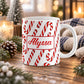 11 oz personalized candy cane pattern mug with red and white candy canes and custom name in red script on both sides, on a wooden table with winter decor around it including pine cones, pine boughs, candles, and a cozy plaid blanket.