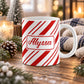 11 oz personalized candy cane stripe mug with red and white diagonal stripes and custom name in red script on both sides, on a wooden table with winter decor around it including pine cones, pine boughs, candles, and a cozy plaid blanket.